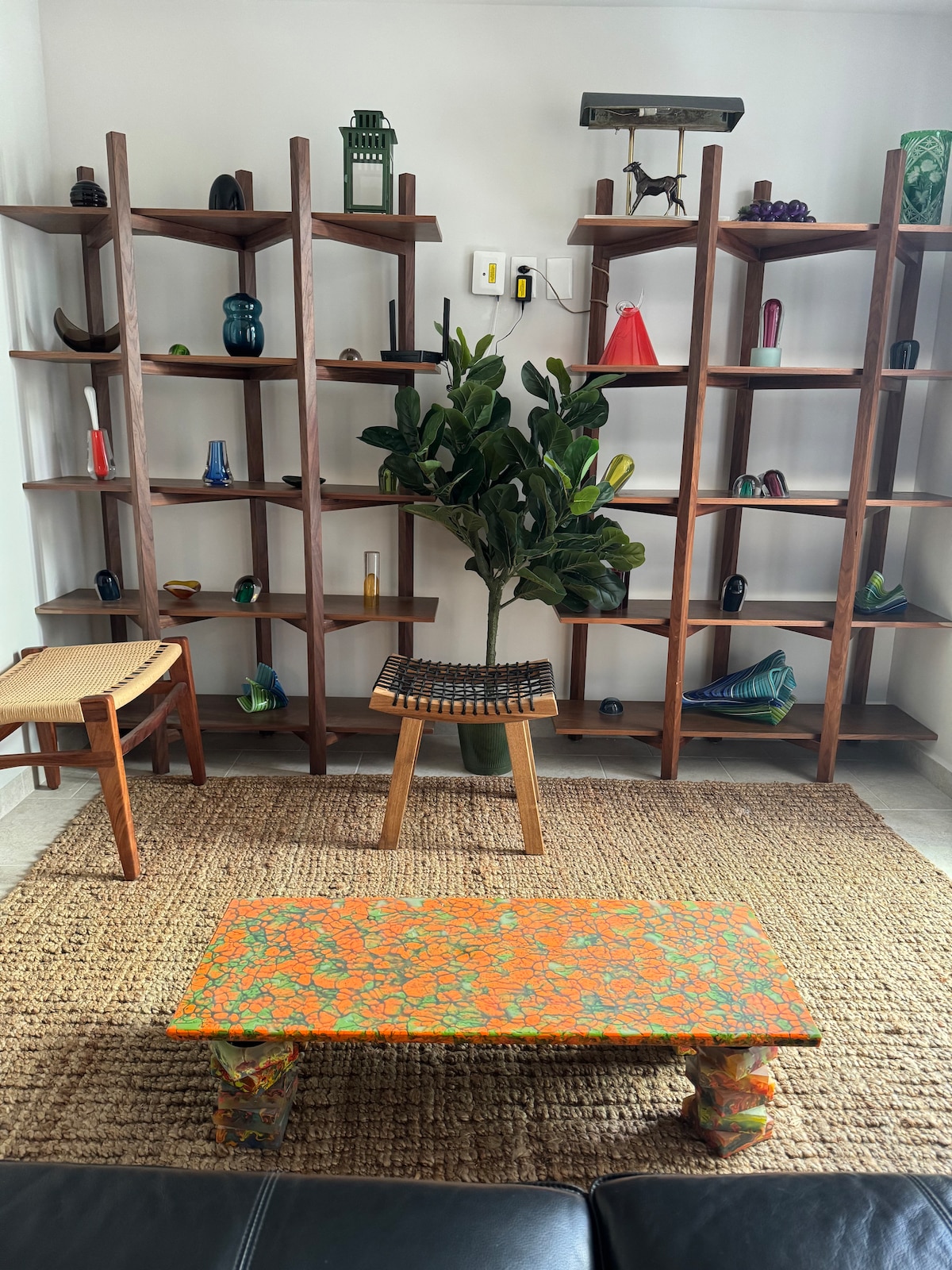 A vibrant seating area features a patterned orange coffee table set on a woven rug. Two open wooden shelving units display an array of colorful decorative items, complemented by a potted plant situated in the center. A simple chair is seen nearby.