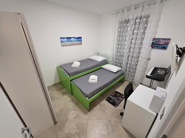 Este é o segundo quarto iluminado, que recebe você com duas camas prontas para oferecer descanso. Na frente, encontramos uma TV e uma mesa arrumada com um piano esplêndido e, finalmente, uma janela francesa com acesso direto ao grande terraço mobilado.