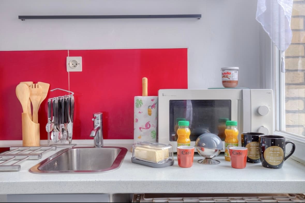 A modern kitchen area features a simple countertop with a stainless steel sink and a red backsplash. Various utensils are neatly stored, and a microwave sits next to colorful mugs and bottles of juice. A butter dish and a globe add functional and decorative elements.
