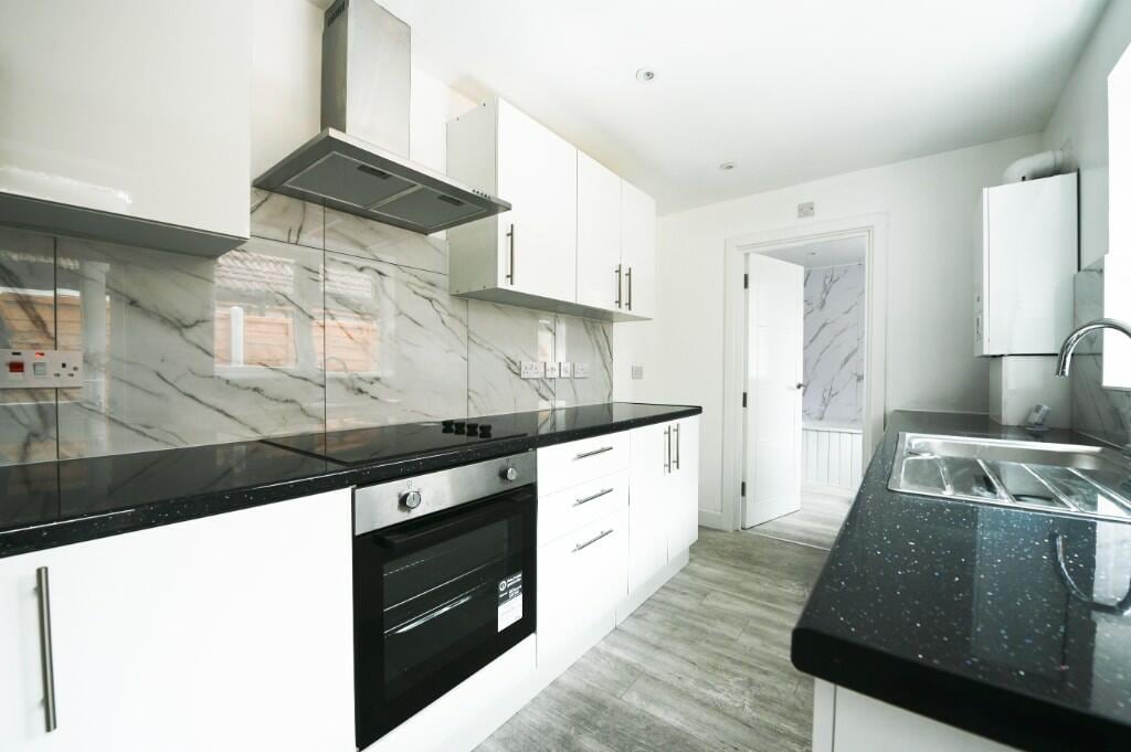 A modern kitchen is depicted, featuring sleek white cabinetry and a dark countertop with subtle flecks. Stainless steel appliances are visible, including an oven and a range hood. The walls showcase marble-patterned tiles, and natural light flows through a nearby door leading to another area.
