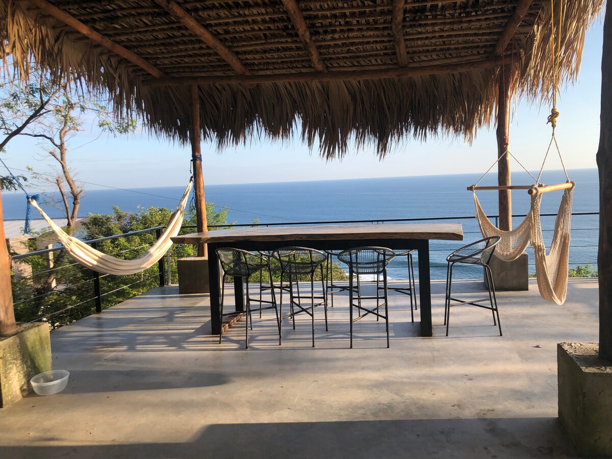 An outdoor dining area features a long wooden table surrounded by black metal chairs, sheltered under a thatched roof. Two hammocks hang nearby, offering relaxed seating options. The expansive ocean view serves as a serene backdrop, enhancing the tranquil atmosphere of this space.