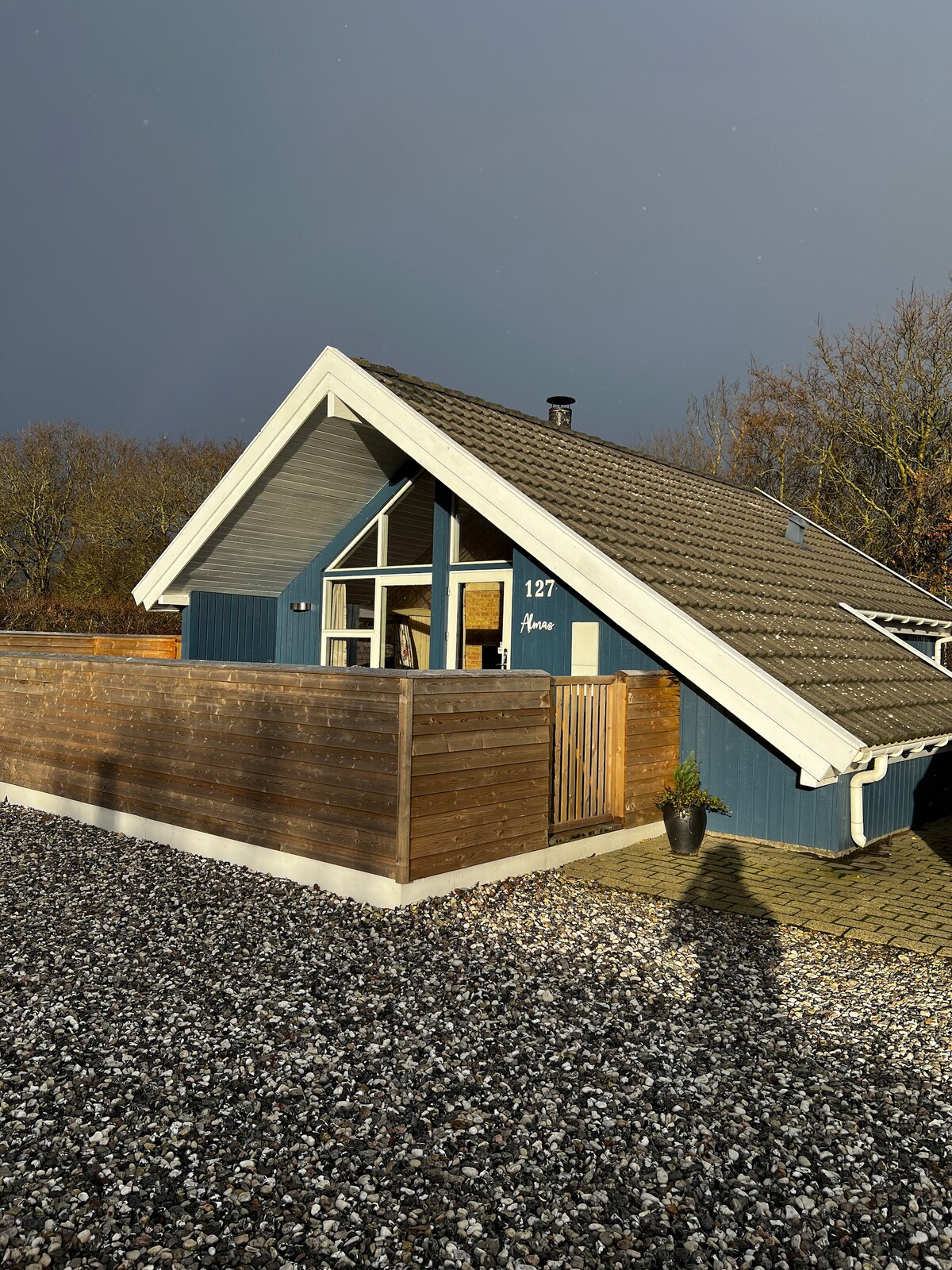 The exterior of a charming blue house is showcased, featuring a sloped roof and wooden accents. The entrance is framed by a wooden fence. Surrounding gravel paths and sunlight filtering through trees create a serene ambiance.