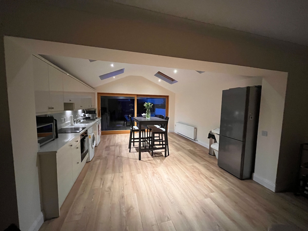 An open-plan kitchen and dining area features modern appliances, including a large refrigerator and a washing machine. A dining table with chairs is positioned centrally, while natural light enters through skylights and a large window, highlighting the warm-toned flooring.