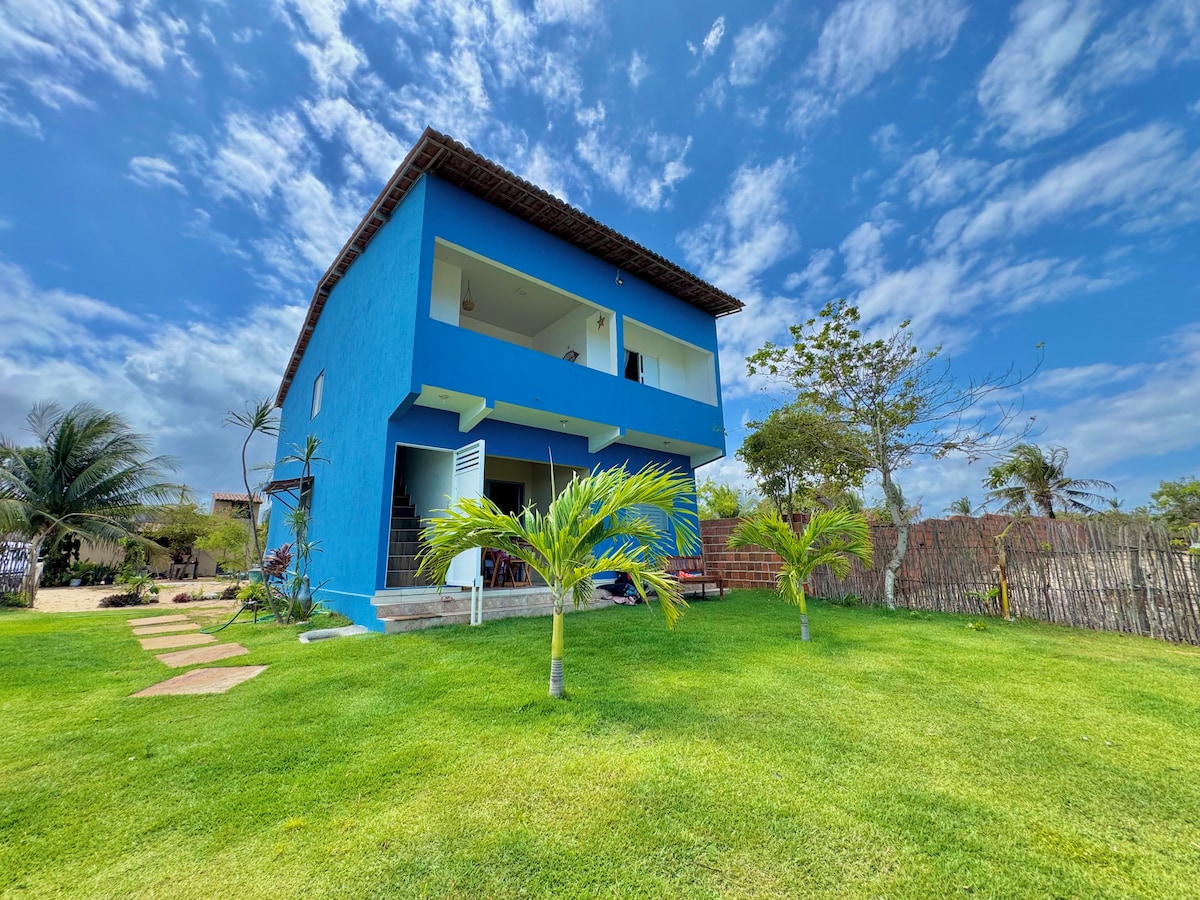 A two-story blue house is set against a clear sky, surrounded by lush green grass and trees. A pathway leads to the house, which features a balcony on the upper level. The outdoor space emphasizes a tranquil and inviting atmosphere.