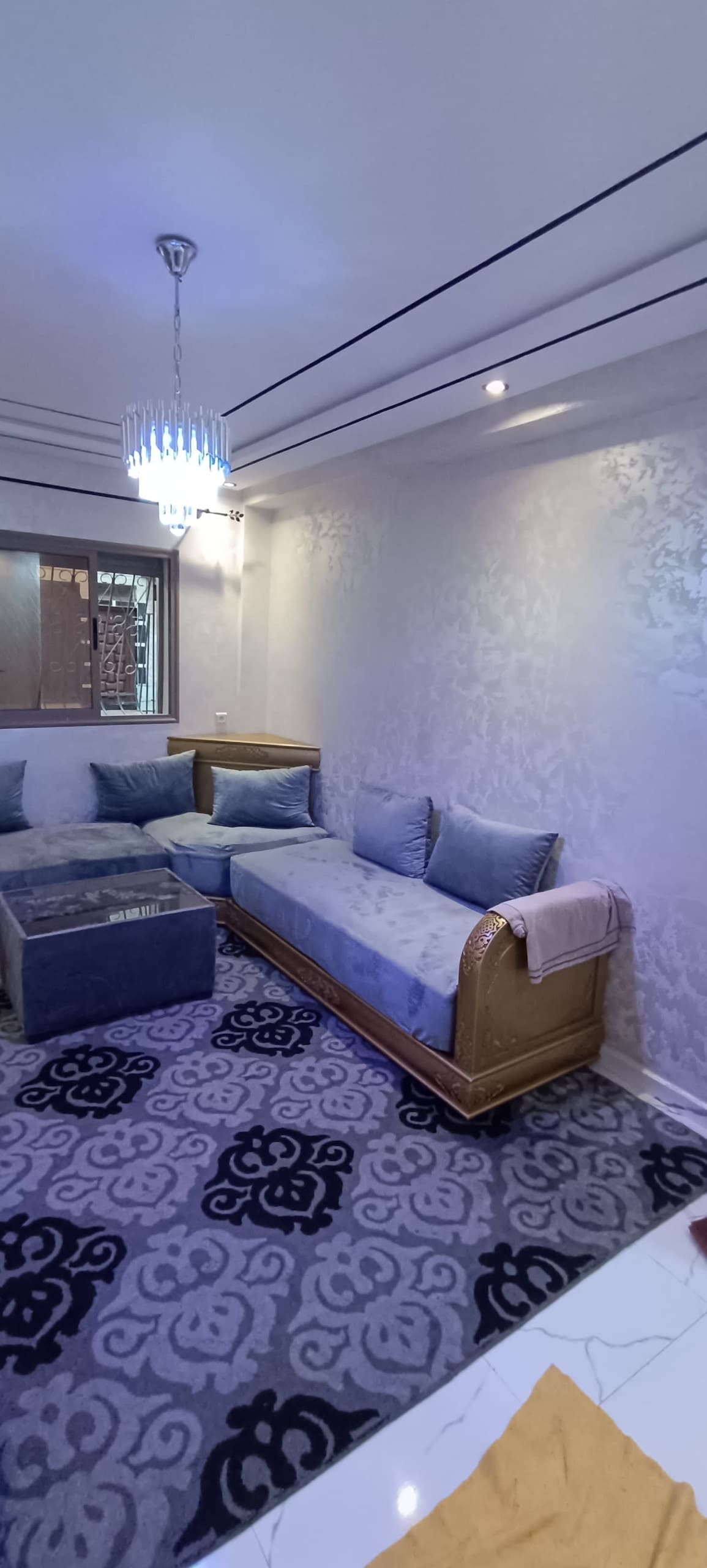 A thoughtfully arranged seating area features a stylish daybed adorned with cushions and a matching ottoman. The space is illuminated by a modern chandelier, complementing the elegant design of the wallpaper and the plush patterned rug underfoot.