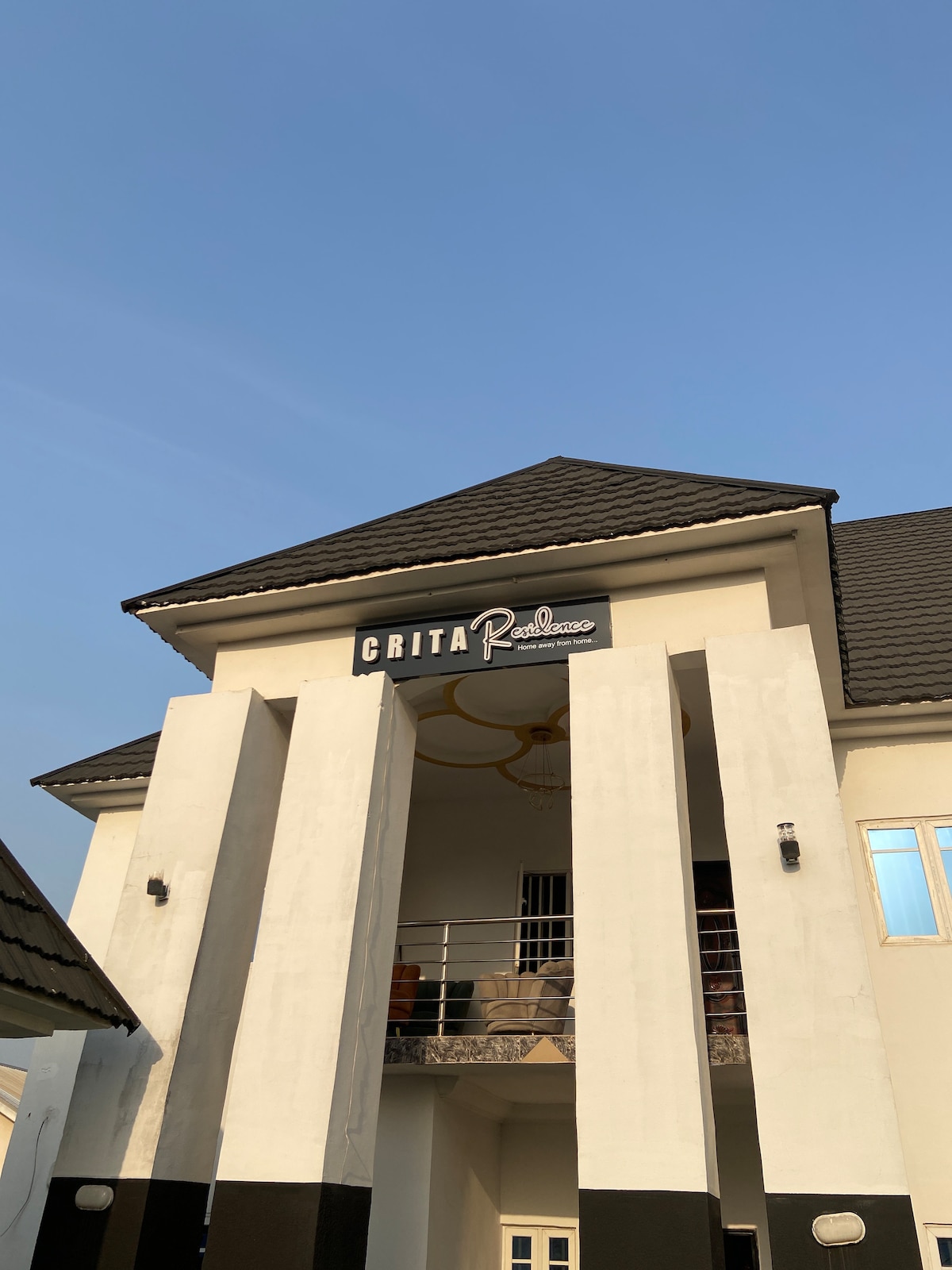 The exterior of the building features a bold sign with the name 'CRITA Residence' prominently displayed. A symmetrical facade is enhanced by a textured roof and a combination of white and dark accents. Windows allow for natural light to brighten the entrance area.