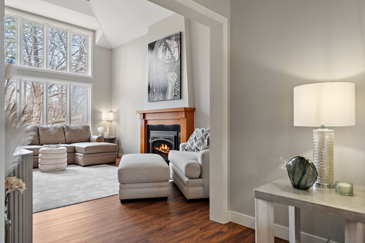 A welcoming living area is bathed in natural light from large windows, highlighting a cozy fireplace surrounded by soft seating. A plush rug adds comfort underfoot, while a stylish lamp provides additional warmth, creating an inviting space for relaxation.
