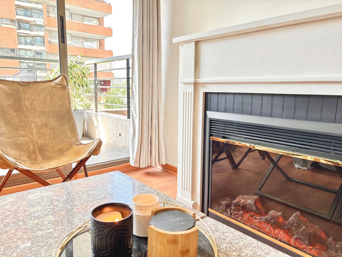 A cozy living space features a comfortable chair with a soft fabric finish, positioned beside a modern fireplace. A marble coffee table holds decorative candles, while natural light fills the room through the glass doors leading to a balcony.