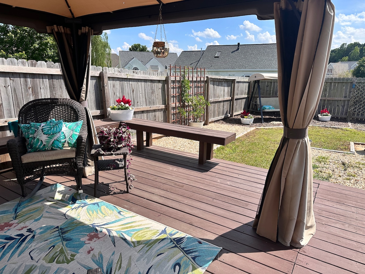 A covered outdoor space is revealed, featuring a comfortable wicker seating area adorned with a colorful cushion. A wooden bench is positioned nearby, and a rug with tropical patterns adds a vibrant touch. Potted flowers enhance the natural setting in the fenced yard.