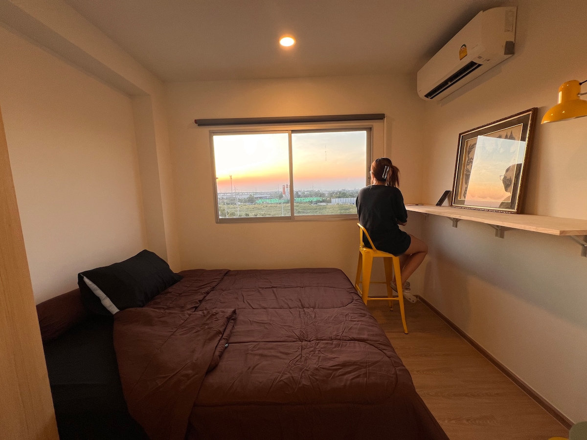 A cozy room is furnished with a spacious bed covered in a brown comforter. A seating area with a yellow chair is positioned by a large window, offering a view of the sunset. An air conditioning unit and wall-mounted mirror are visible.