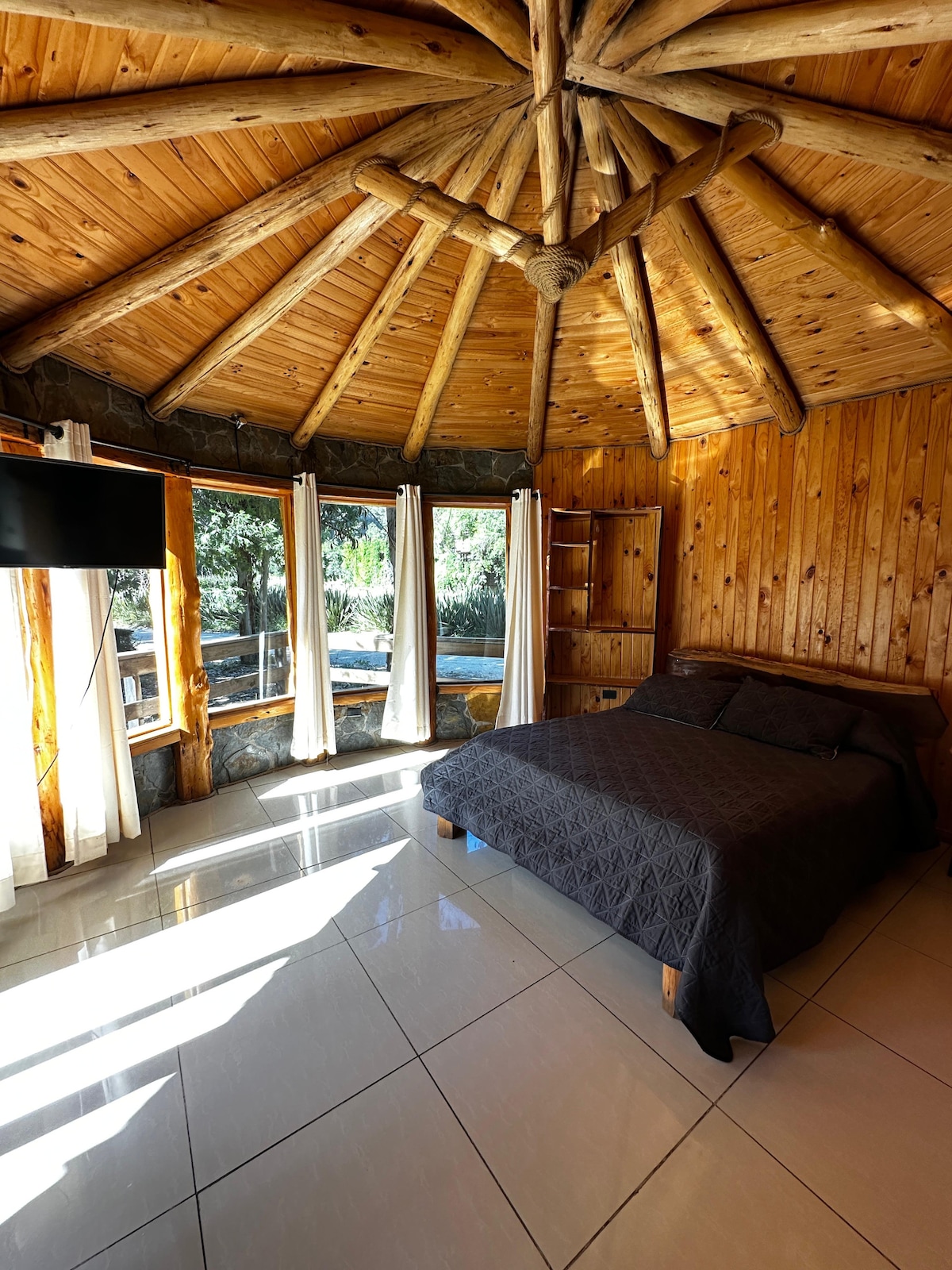 A circular room features a rustic wooden ceiling with exposed beams. A spacious bed is dressed in a dark blanket. Large windows allow natural light to fill the space, while a flat-screen TV is mounted on the wall. Light-colored tiles enhance the room's brightness.