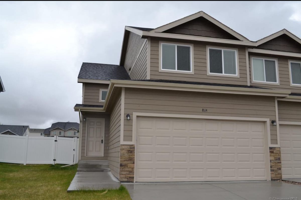 A two-story home exterior is shown, featuring a light brown facade with stone accents. The garage door is designed with multiple panels, and a covered entryway is visible. A spacious lawn leads to a white fenced area, creating an open and inviting atmosphere.