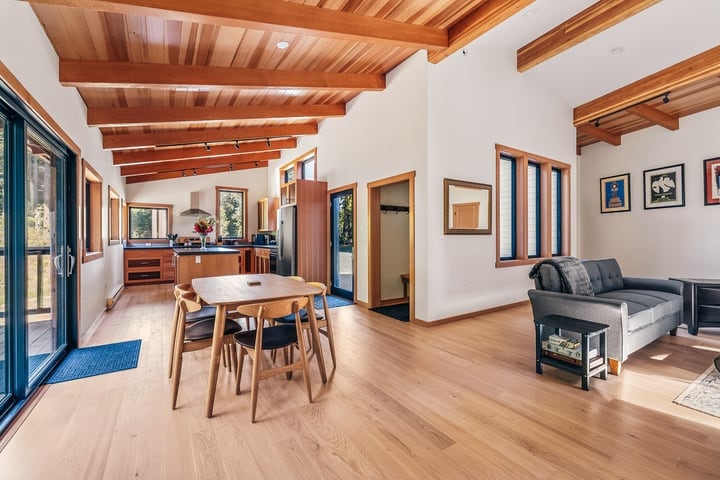 A spacious living area features warm wooden beams and a light wood floor. A comfortable gray sofa is positioned near a large window, alongside a simple dining table with chairs. The open design connects the living space to a modern kitchen area in the background.