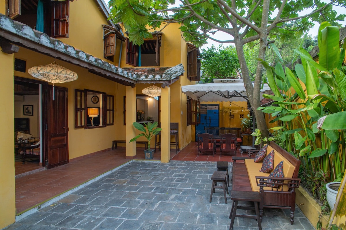 An inviting outdoor courtyard is framed by vibrant greenery and tropical plants. Traditional lanterns hang above a stone pathway leading to wooden seating arrangements, which are comfortably set under the shade. Bright windows of the nearby structure enhance the area’s natural light.