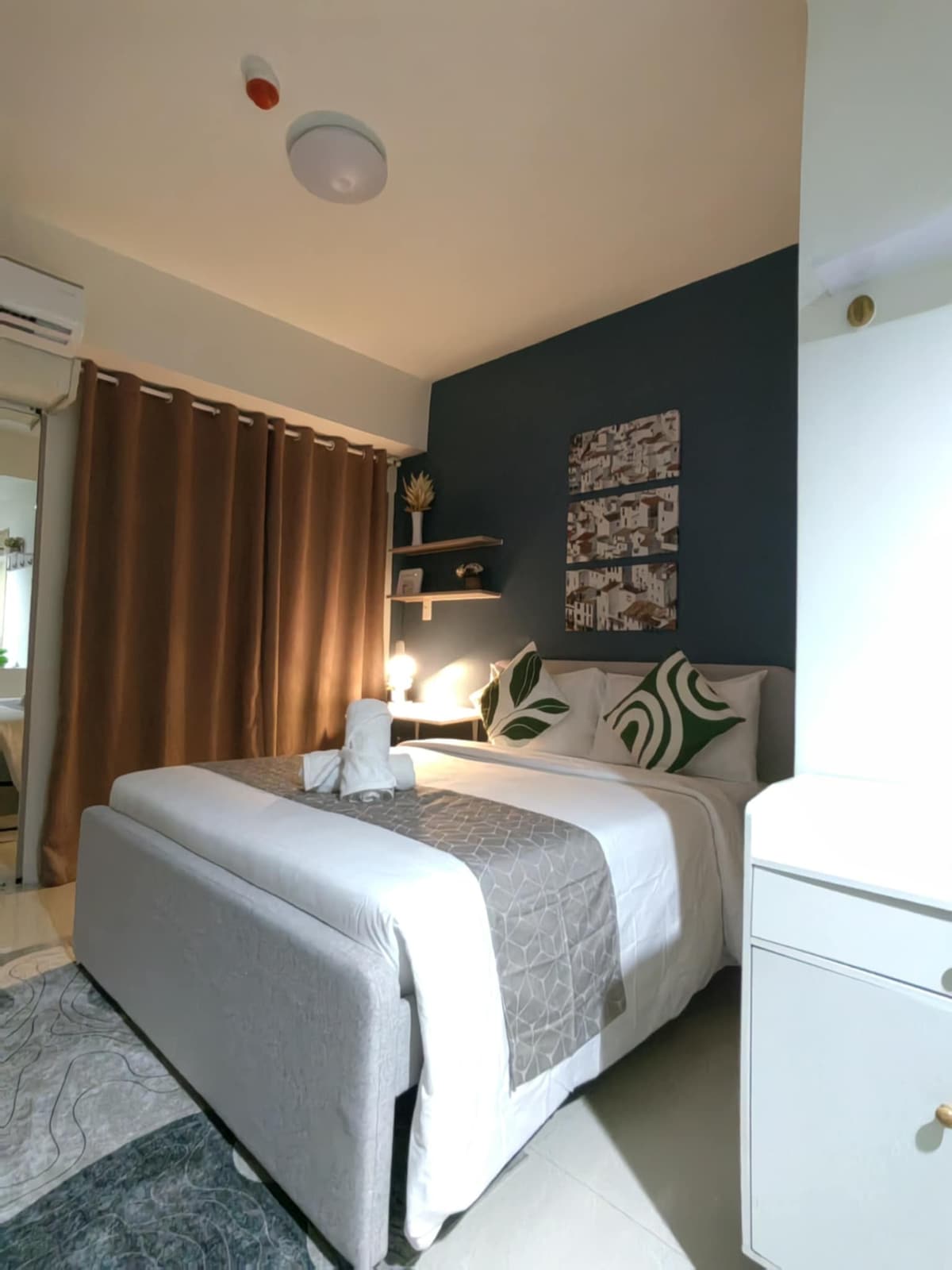 A cozy bedroom area features a spacious bed dressed in neutral tones with decorative green pillows. A dark accent wall adds depth, while soft curtains frame the window. A small shelf with plants and an artwork piece enhances the room's inviting decor.