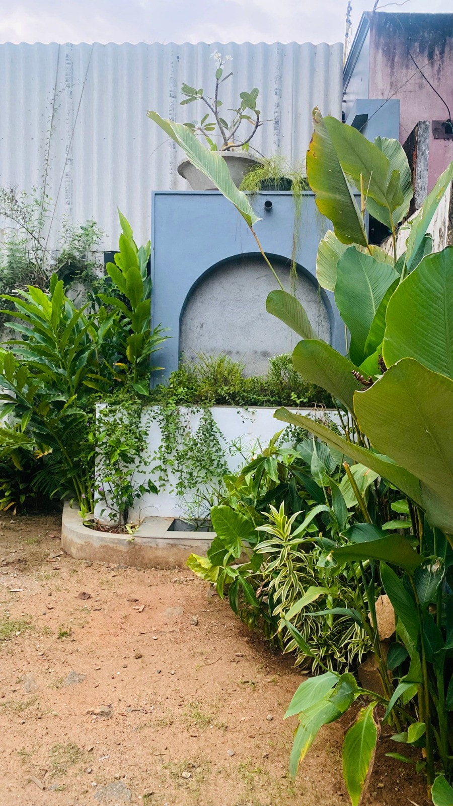 A landscaped area is surrounded by lush green plants, featuring soft earth tones. A concrete structure with a rounded archway is partially hidden among foliage, creating a serene and inviting ambiance in the outdoor space.