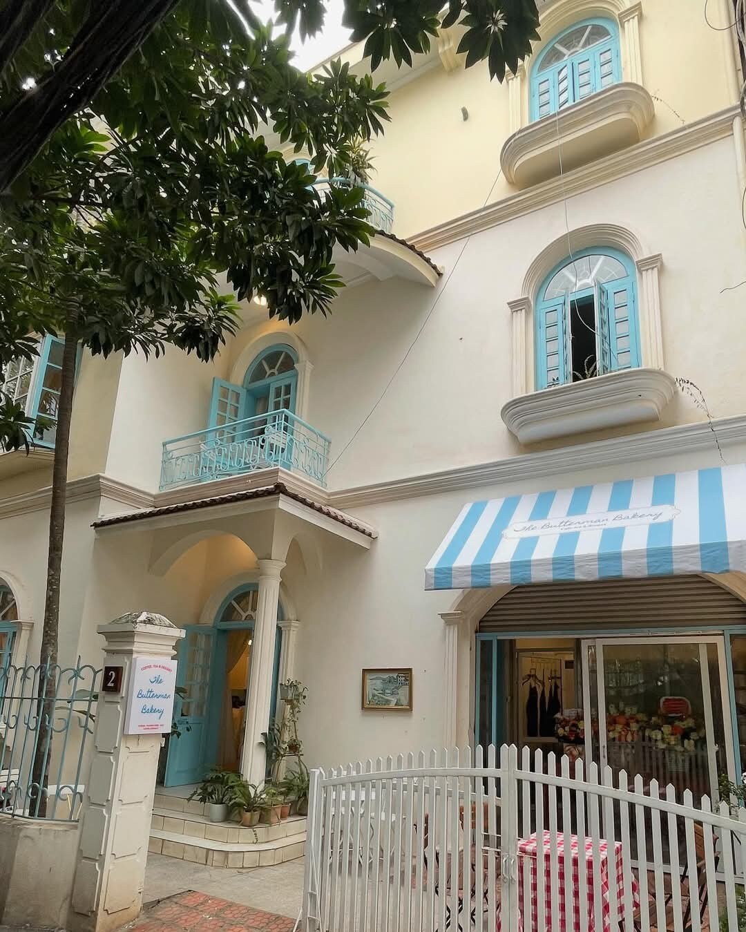 The exterior of a French-style villa is displayed, showcasing light-colored walls and distinctive blue shutters. A striped awning provides coverage over the entrance, while a white picket fence surrounds the property. Lush greenery adds a natural element to the inviting atmosphere.