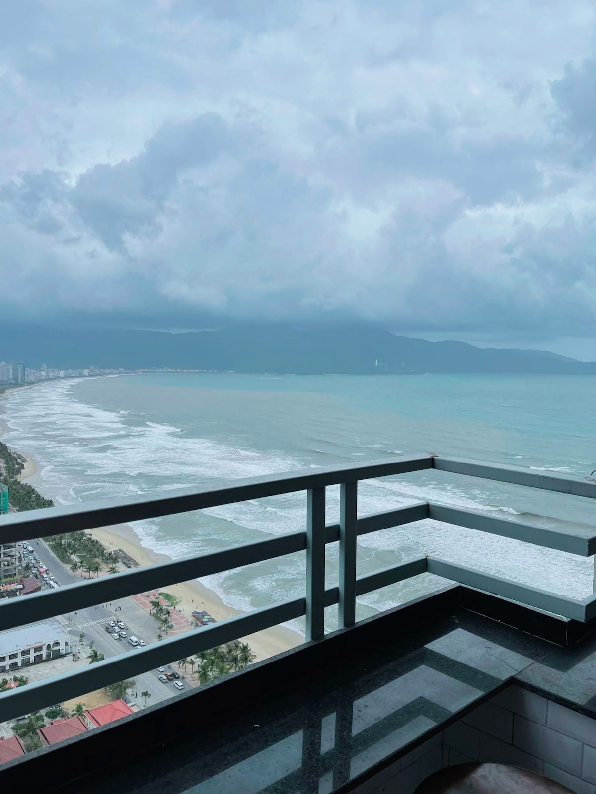 A sweeping view of the coastline is captured from a high balcony, showcasing gentle waves lapping at the shore. The expansive ocean stretches towards distant mountains under an overcast sky, while city buildings are visible along the horizon.