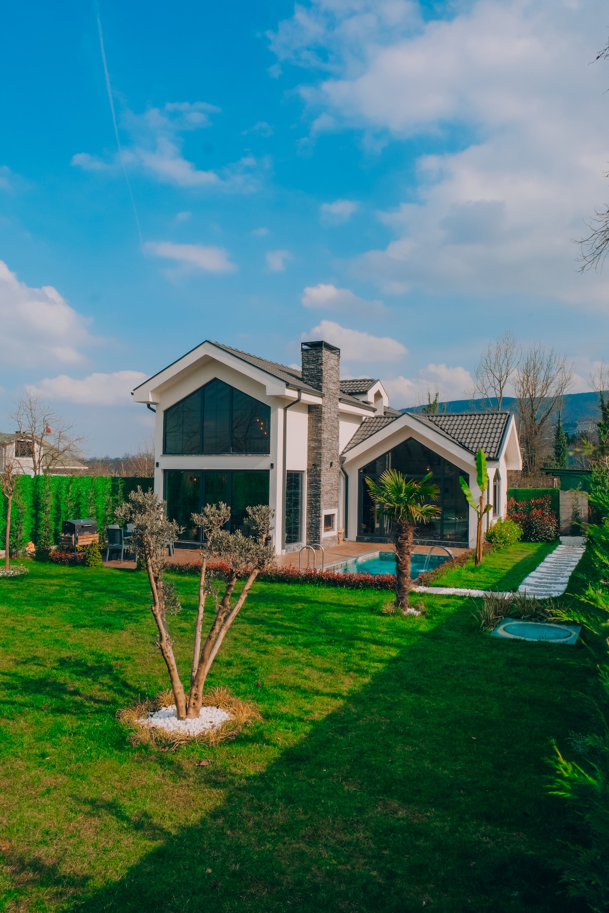 The villa's exterior showcases a contemporary design with large glass windows, allowing natural light to fill the space. A well-maintained garden surrounds the property, featuring manicured grass and small trees, contributing to a serene outdoor environment.