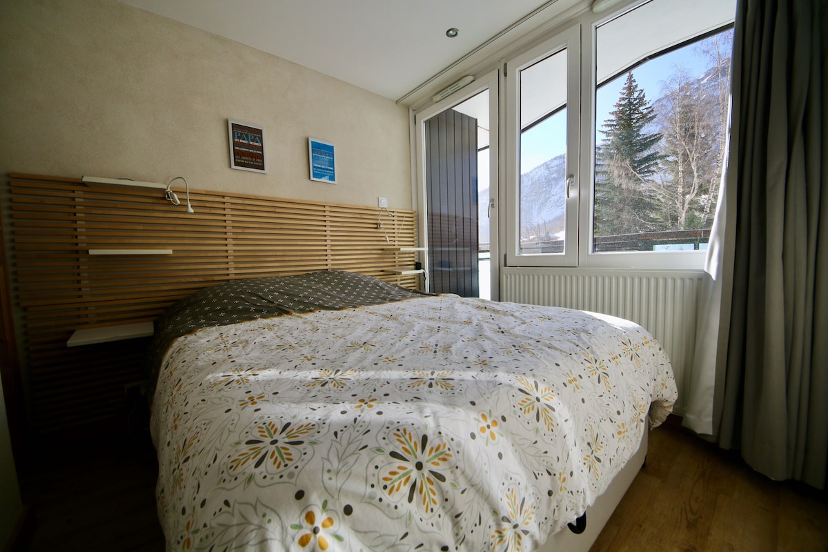 A bright bedroom features a spacious bed with a floral-patterned duvet cover. Large windows provide natural light and showcase a view of the surrounding trees and mountains. Soft curtains frame the windows, enhancing the serene atmosphere.