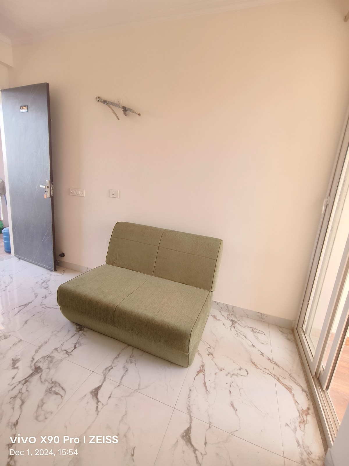 A simple green lounge chair is positioned against a light-colored wall in a bright room. The glossy marble floor reflects natural light coming through a nearby glass door, enhancing the spacious feel of the area.