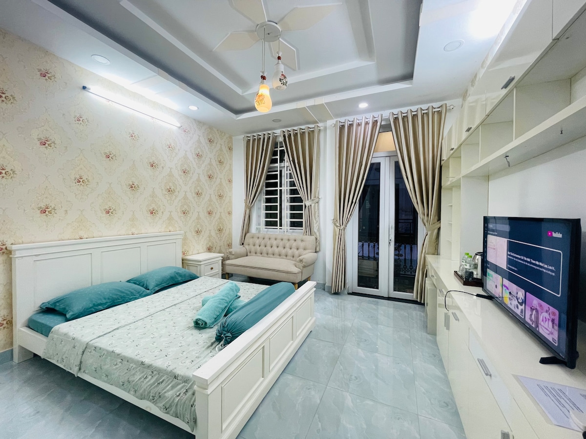 A spacious room features a large bed adorned with teal pillows and a light-colored bedspread. A beige sofa is positioned near the window, which is dressed with elegant curtains. A television is mounted on the wall, and ambient lighting enhances the warm decor.
