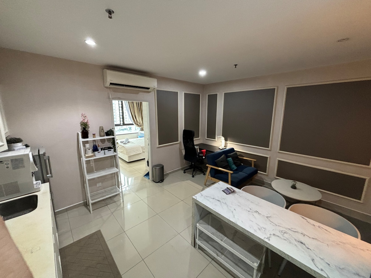 A contemporary living area is shown, featuring a blue sofa and two circular coffee tables. A small dining table is present, complemented by a kitchenette that includes a microwave and a waste bin. Light-colored walls and large windows enhance the spaciousness of the room.