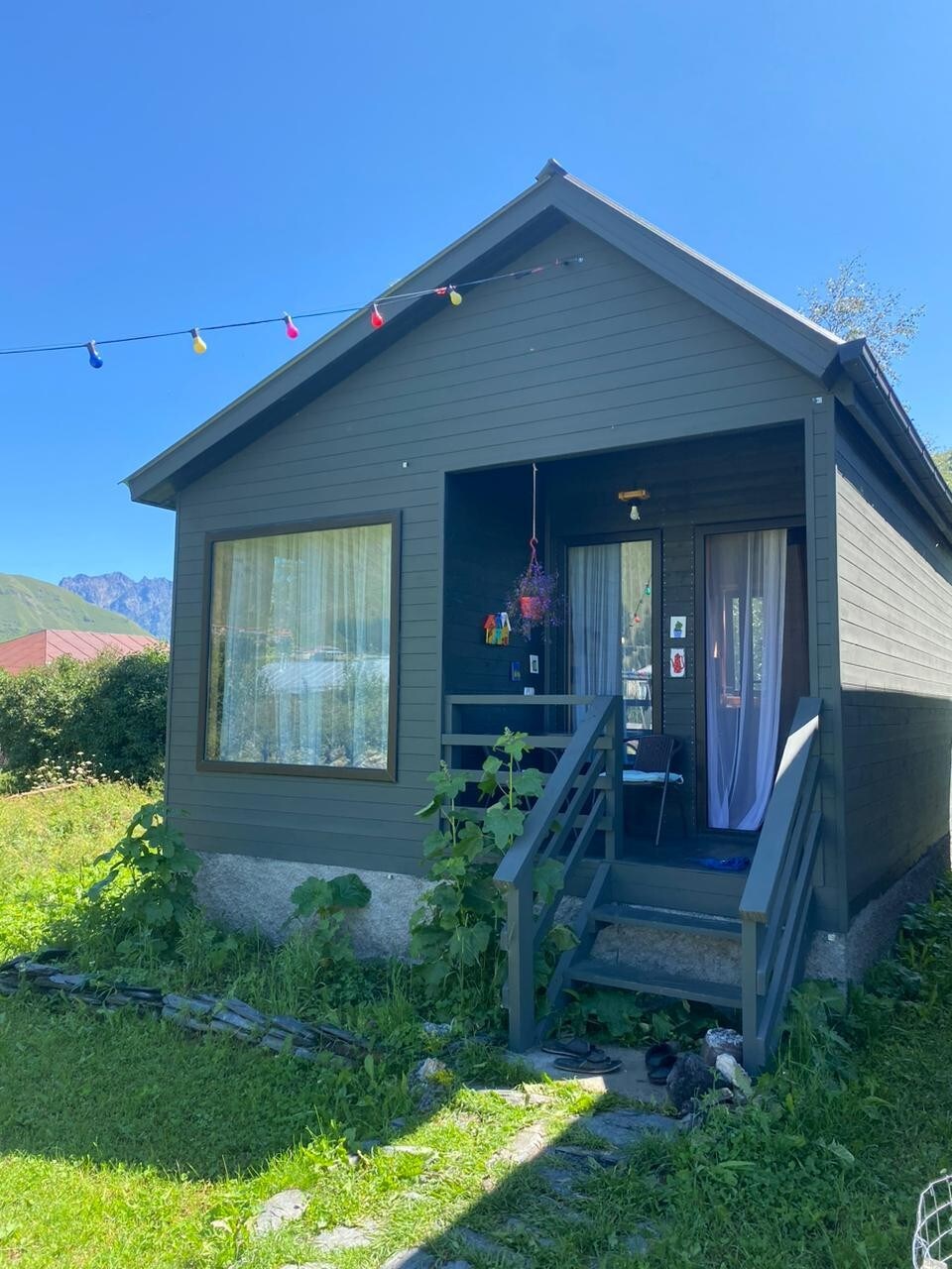 A quaint cabin features a dark wood exterior with a welcoming entryway. Large windows allow natural light to illuminate the space. Colorful lights are draped across the front, enhancing the outdoor area surrounded by greenery.