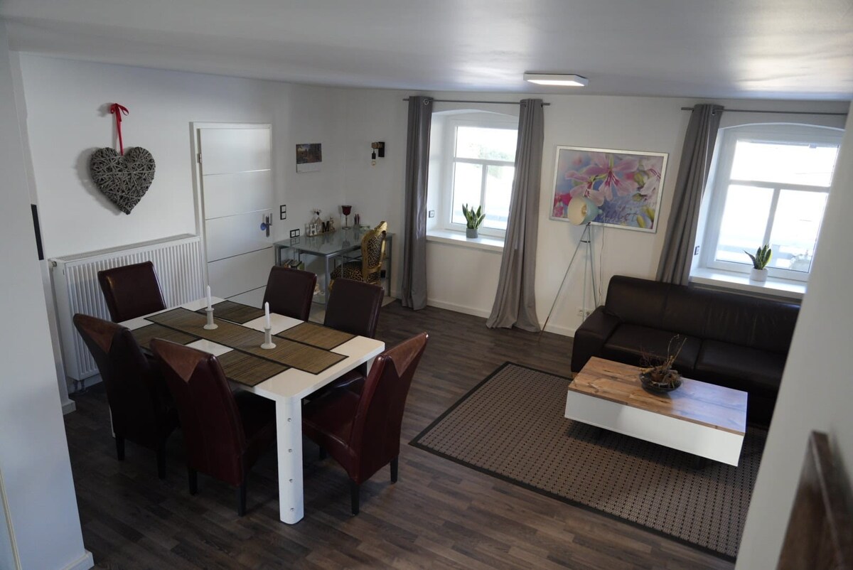 A spacious living area features a dining table set for six, framed by dark-wood chairs. Light-colored walls are complemented by large windows, enhancing brightness. A comfortable couch sits opposite a decorative coffee table, with a wall adorned by artwork and a heart-shaped decor piece.