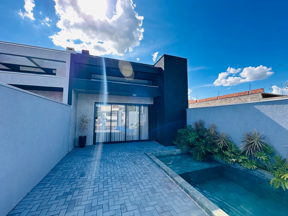A modern exterior is showcased under a bright blue sky, featuring a sleek design with large glass sliding doors. The outdoor space includes a small pool surrounded by lush greenery and paved stone flooring, providing a serene atmosphere.