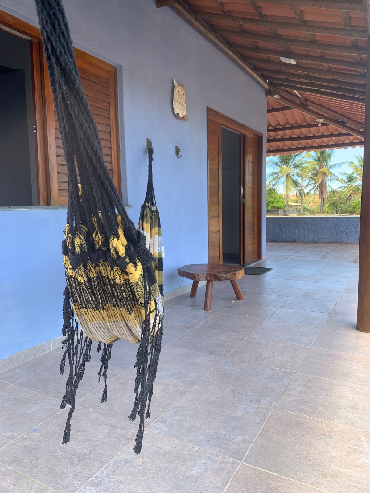 A stylish hammock hangs from a support beam, complemented by a small wooden stool nearby. The tiled floor extends towards an open doorway, revealing a view of palm trees outside, enhancing the sense of tranquility in this outdoor space.