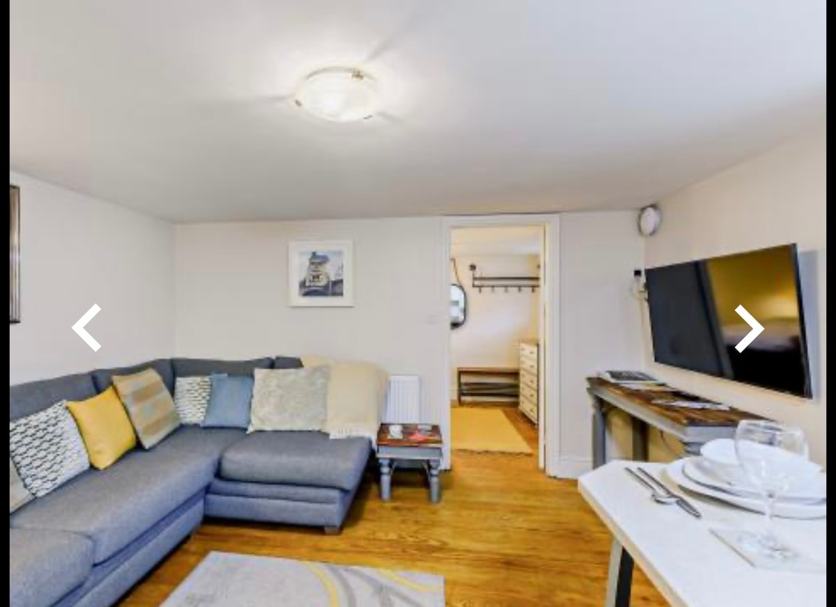 A comfortable living area is highlighted by a spacious sectional sofa adorned with decorative cushions. A flat-screen television is mounted on the wall, opposite a small dining table set for four. Light wooden flooring extends throughout the space, enhancing the cozy ambiance.