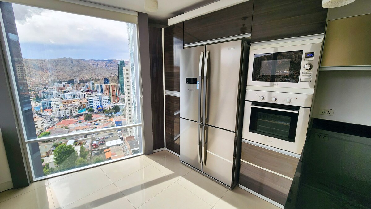 The kitchen is equipped with modern stainless steel appliances, including a refrigerator, oven, and microwave, positioned against a sleek dark cabinetry backdrop. A large window offers expansive city views, while natural light fills the space, highlighting the clean lines of the design.