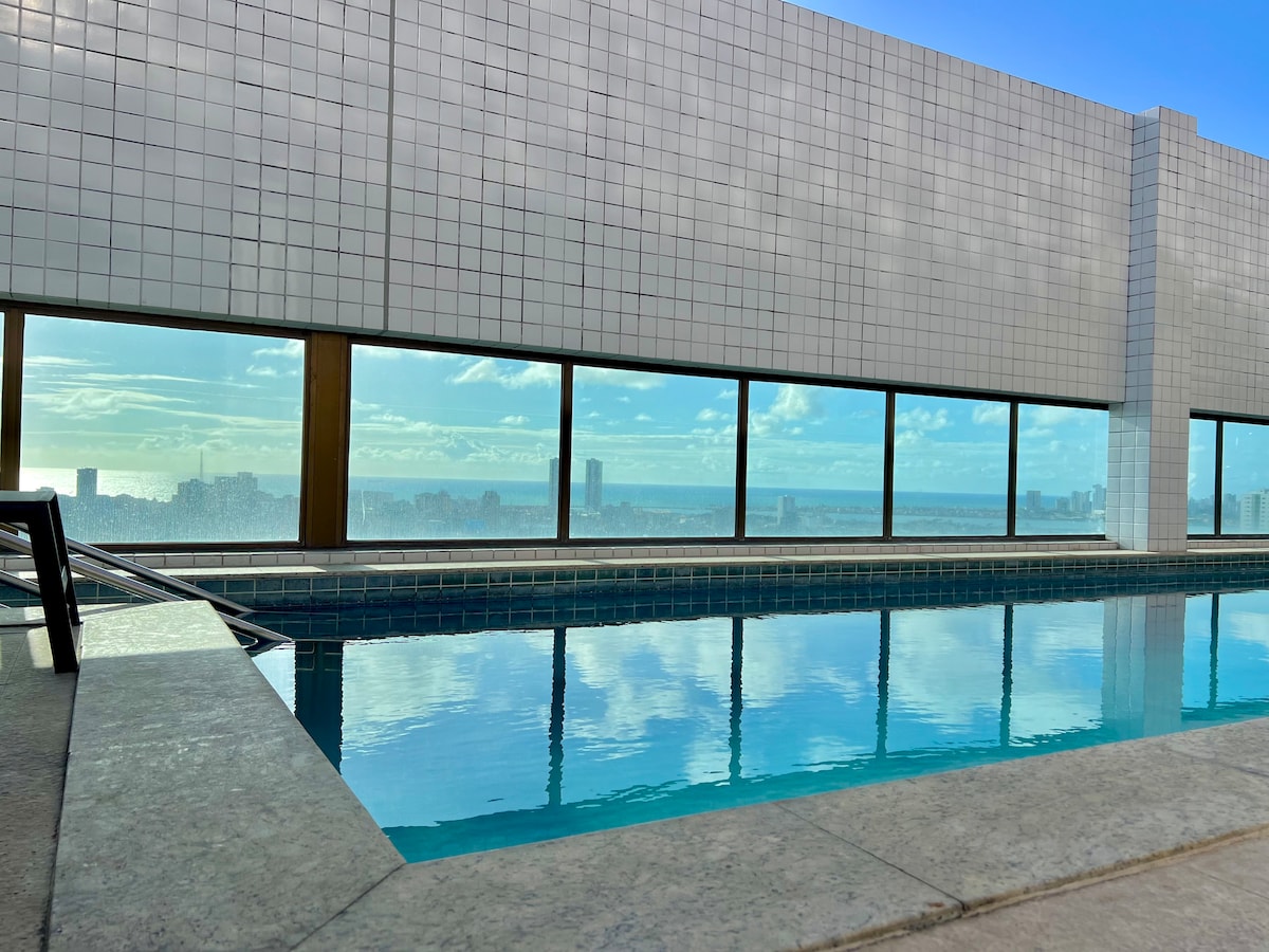 An indoor pool reflects the scenic view through large glass windows. The serene water contrasts with the sleek tile surrounding, creating a calming ambiance. The skyline can be seen in the background under a clear blue sky, enhancing the spacious feel of the area.