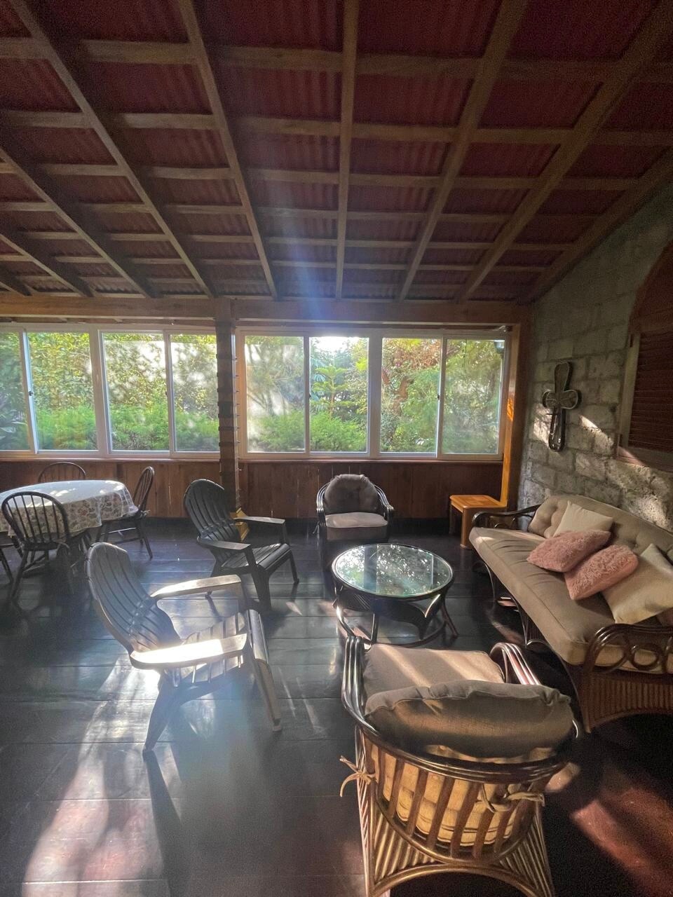 A sunlit seating area features a mix of wicker chairs and a glass-top table, surrounded by large windows that frame the lush greenery outside. A cozy sofa provides additional seating, creating a welcoming space for relaxation.