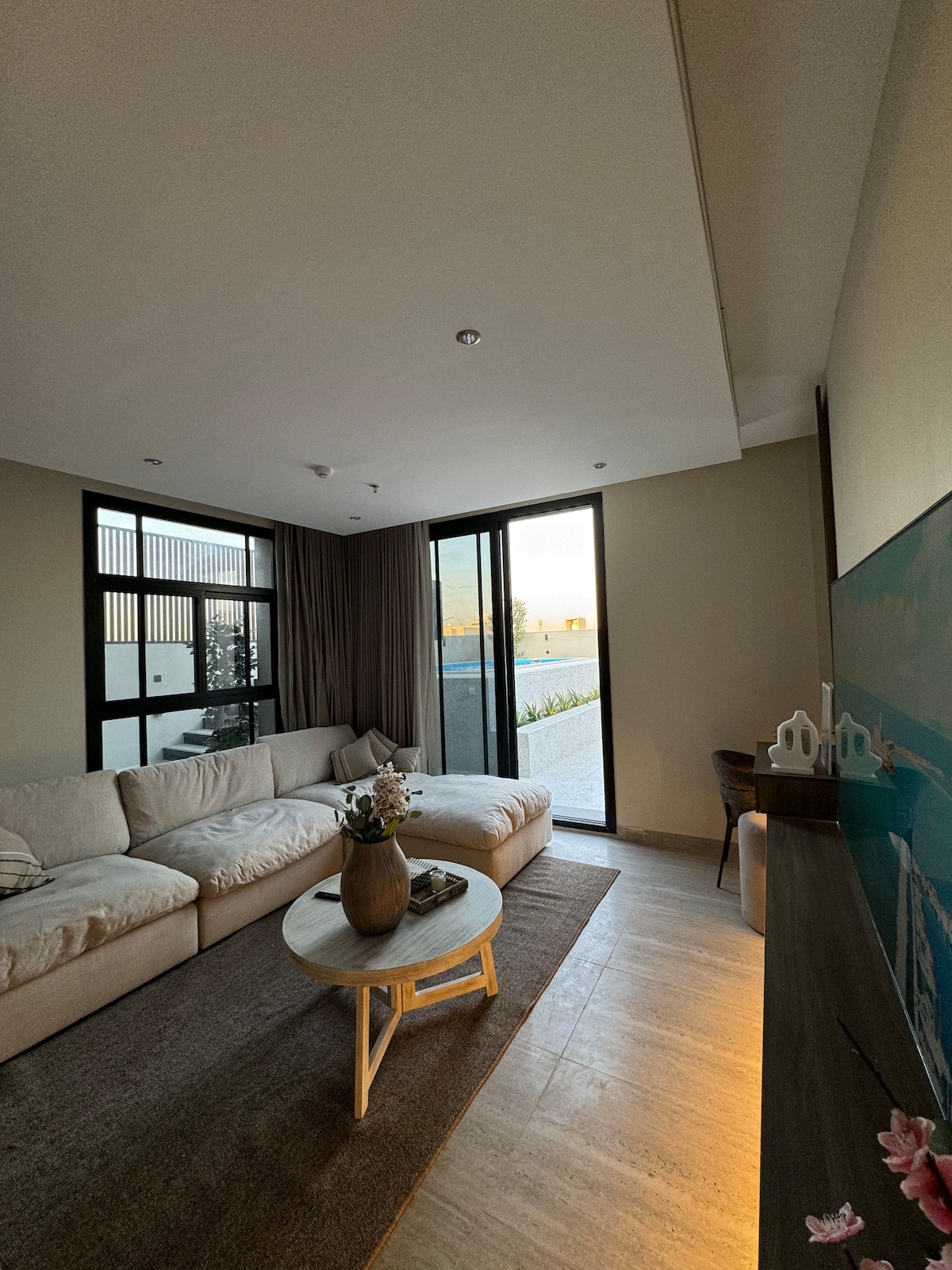 A modern living area features a spacious light-colored sectional sofa, accompanied by a round wooden coffee table. Large glass doors allow for ample natural light and provide access to an outdoor space, while a decorative painting adds a subtle artistic touch to the wall.