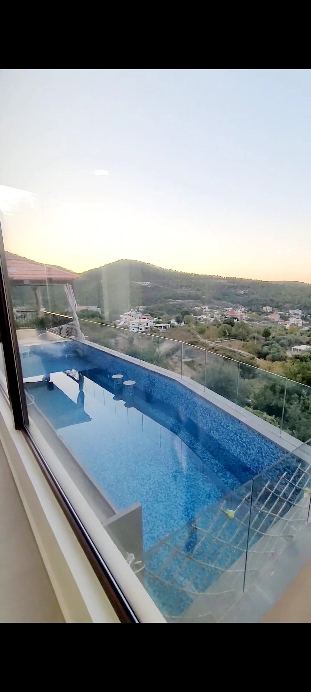 A sleek infinity pool extends from the edge of the balcony, showcasing shimmering blue water. Below, the surrounding hills and distant houses are visible, bathed in warm sunset light. The glass railing emphasizes the unobstructed view of the natural landscape.