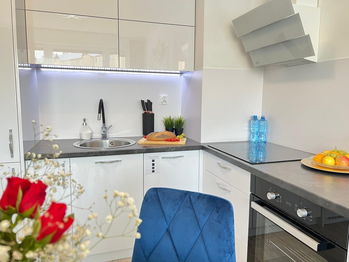 The kitchen area features modern cabinetry with a compact countertop. A small sink is paired with a sleek faucet. A cutting board with food preparation items is visible, along with two bottles of water and a bowl of fruit. Soft lighting enhances the space.