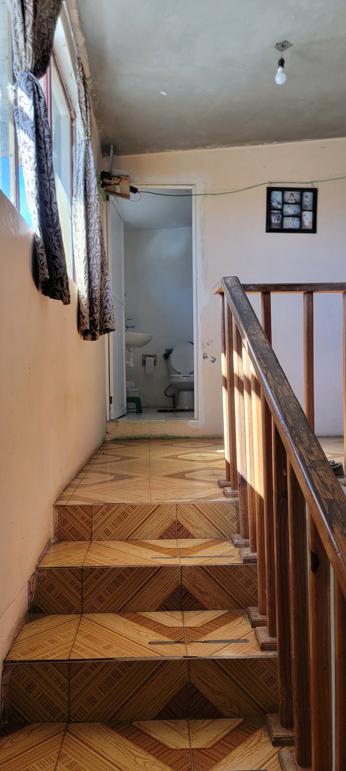 A staircase with wooden handrails leads to a brightly lit landing. Natural light enters through a window adorned with patterned curtains. A door at the end of the hallway is partially visible, indicating access to a bathroom, while framed photos are displayed on the wall.