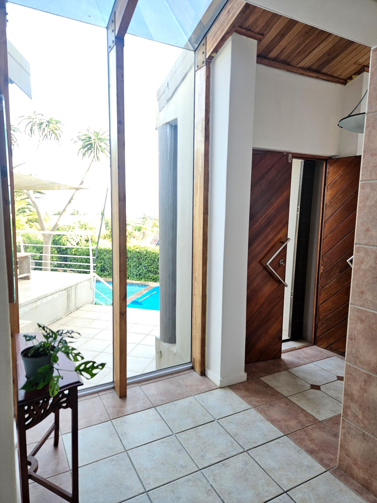 An entryway features large glass doors that provide a view of the outdoor pool area. Natural light illuminates the space, highlighting the tiled floor. Wooden accents and a small plant add a touch of warmth to the modern design.