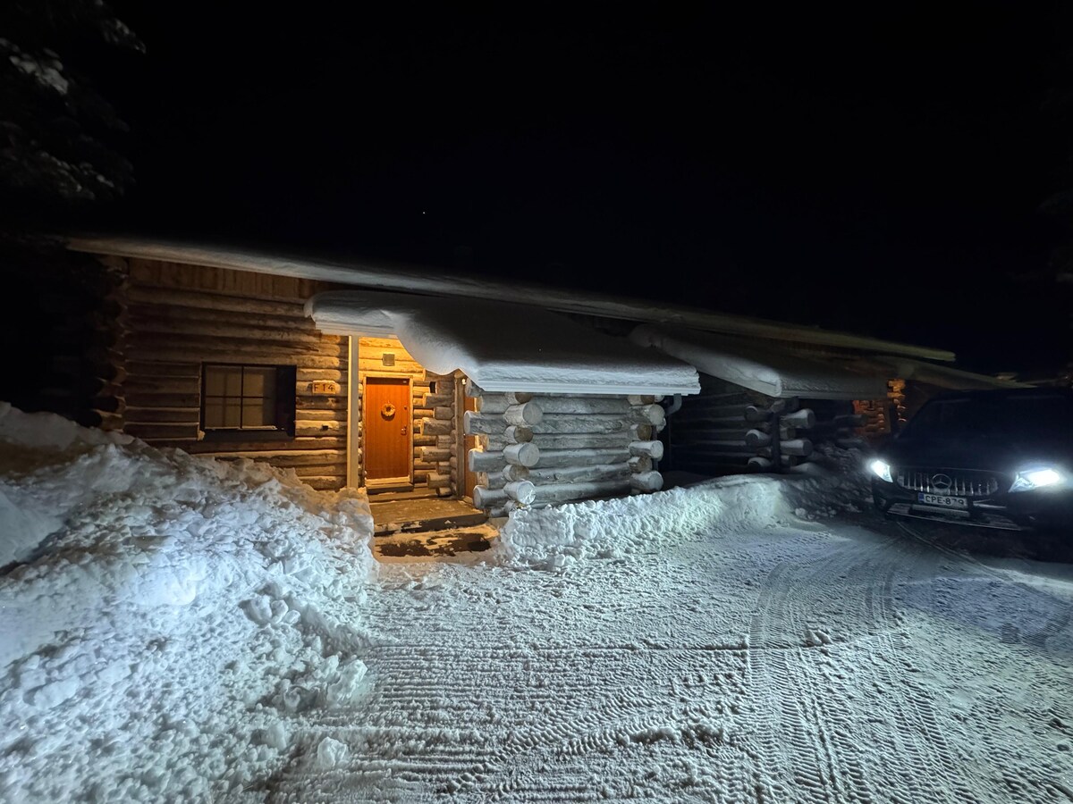 A rustic log cabin is illuminated by warm light from the entrance, surrounded by a blanket of snow. Car headlights cast a soft glow on the driveway, and the eaves are adorned with a thick layer of snow, enhancing the winter scene.