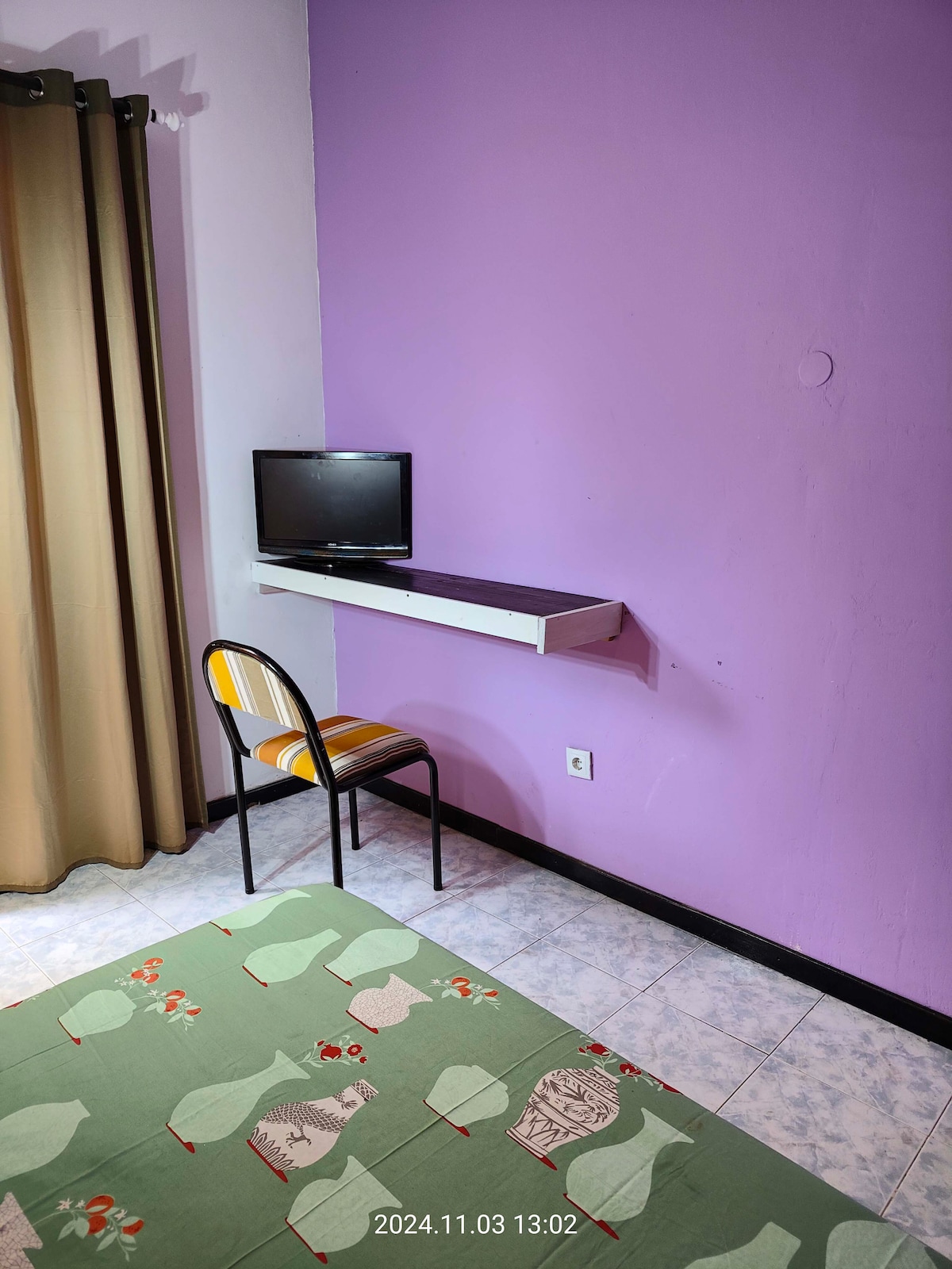 A compact space features a small wall-mounted television above a simple table. A chair with a striped pattern is positioned beside the table. The wall is painted in a soft purple, complemented by light brown curtains that partially cover a window.