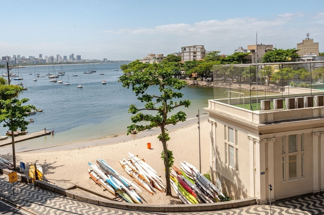 A view of the waterfront features a sandy beach framed by a tree. Several colorful paddleboards are lined up on the shore. In the distance, calm waters are dotted with sailboats, with the city's skyline visible on the horizon.
