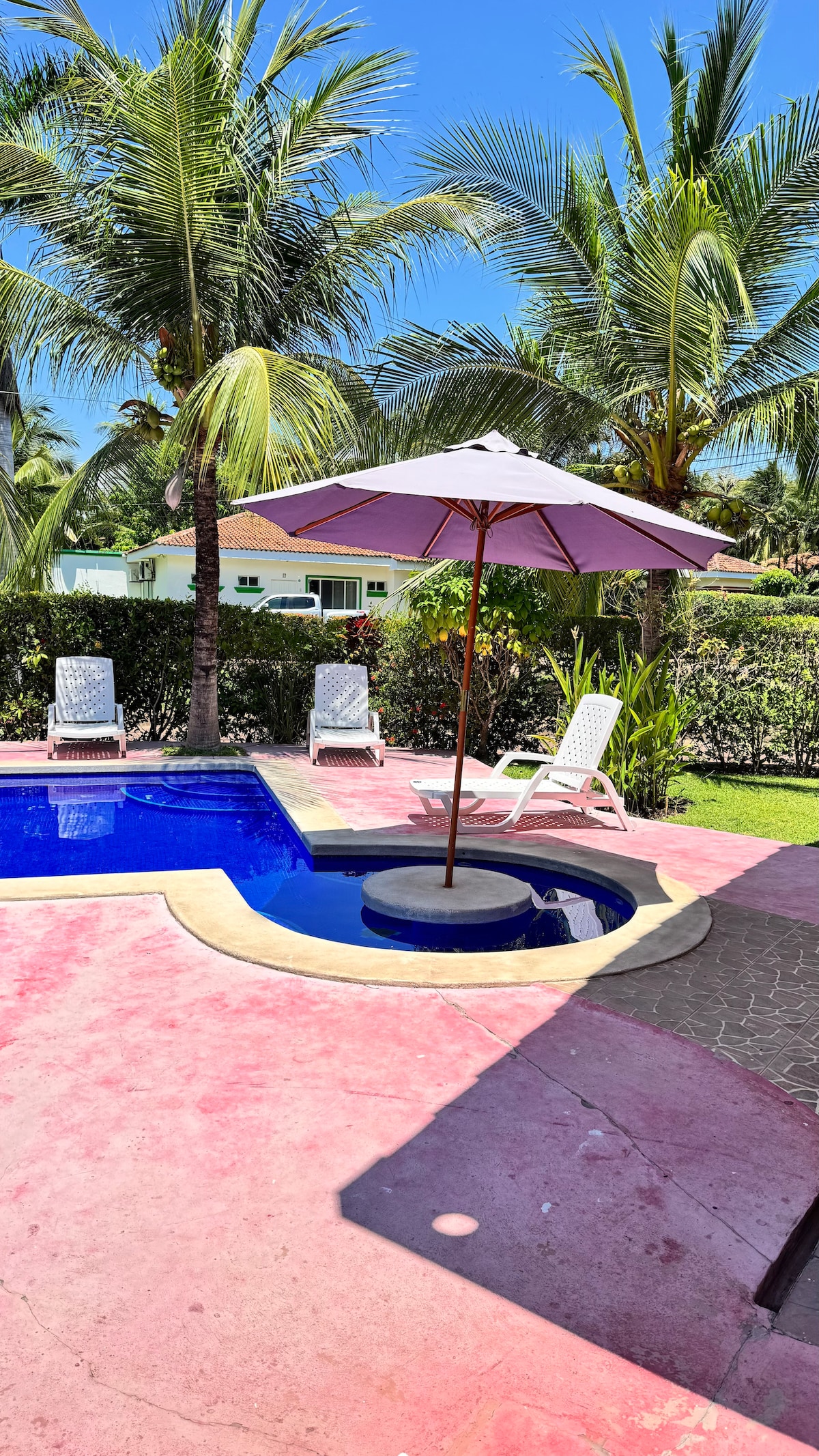 A private pool area is shown, featuring a circular hot tub surrounded by a tropical backdrop. A pink umbrella provides shade over the hot tub, while two white lounge chairs sit beside the pool. Lush greenery enhances the overall relaxing ambiance.