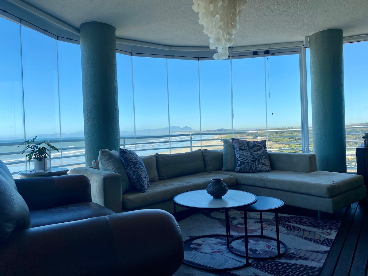 A spacious living area features large windows that offer panoramic views of the ocean and mountains. A comfortable sectional sofa and a round coffee table are present, while a stylish chandelier hangs from the ceiling. A decorative plant adds a touch of greenery to the space.