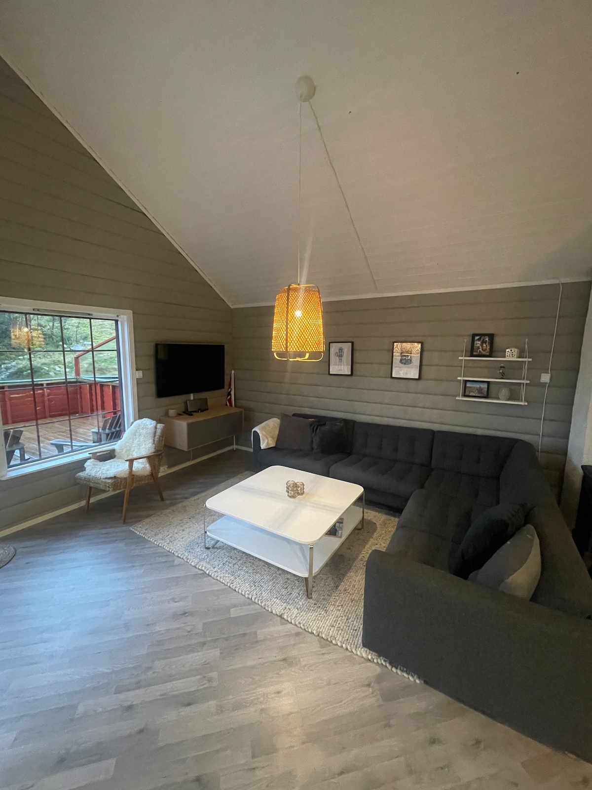 A spacious living area features high ceilings and a cozy gray sectional sofa, complemented by a white coffee table. A modern light fixture hangs above, while a wall-mounted television and framed artwork add character. Large windows provide a view of the outdoors, inviting natural light.