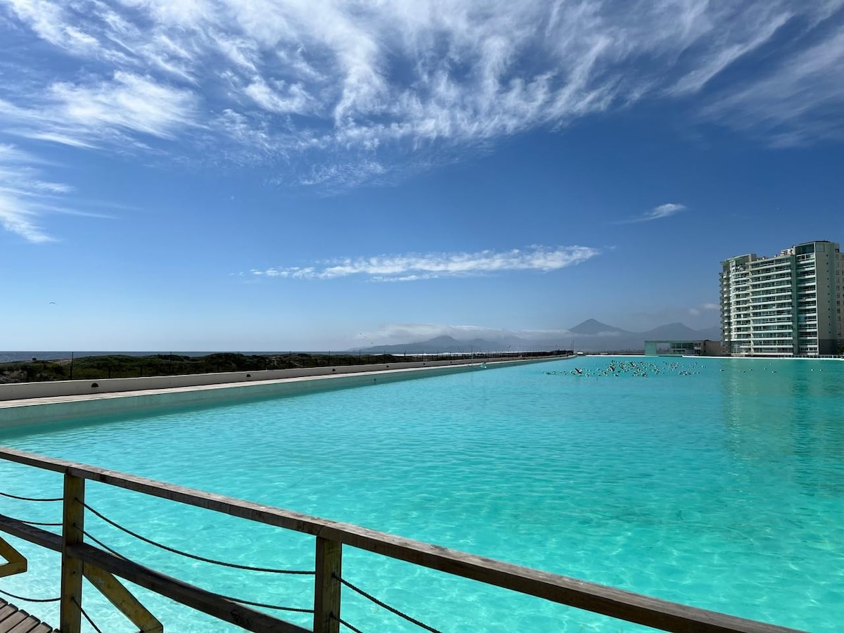 A vibrant, crystal-clear lagoon is surrounded by a wooden deck, reflecting the blue sky and distant mountains. The water appears inviting, with gentle waves and an expansive shoreline visible beneath a bright, sunny atmosphere.