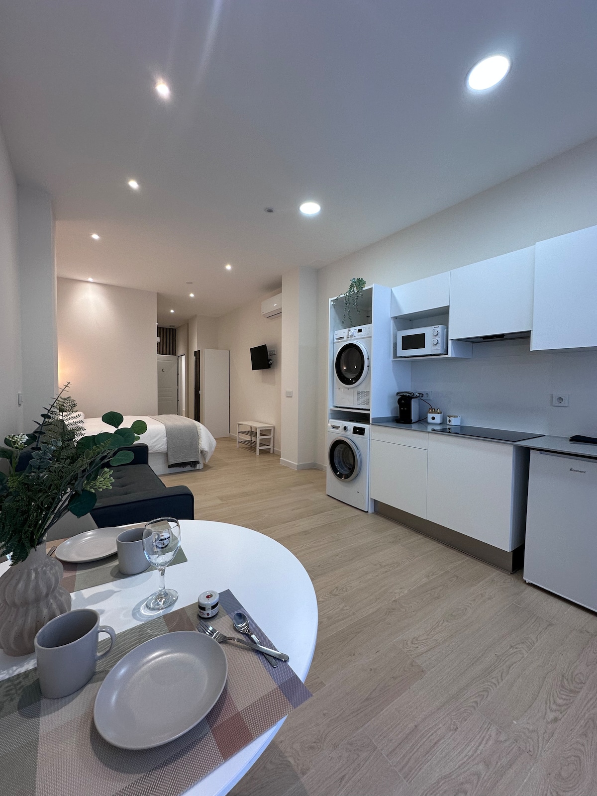An open kitchen is positioned next to a dining area featuring a round table set with two plates and cups. Modern appliances, including a washer and dryer, are integrated into the sleek cabinetry. The spacious layout leads to a cozy sleeping area visible in the background.