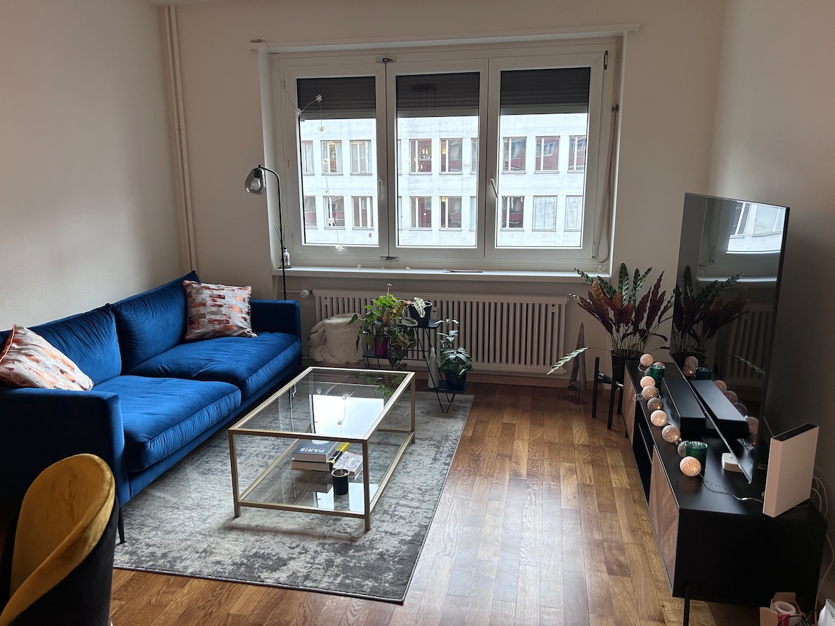A modern living area features a blue sofa adorned with cushions. A glass coffee table sits on a patterned rug, while a large television rests against a white wall. Natural light enters through three large windows, brightening the space.