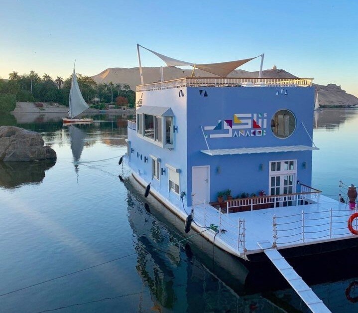 A unique floating structure is illustrated on calm waters, featuring a bright blue exterior. A large deck with seating is visible, complemented by a sunshade. In the background, lush greenery and a traditional sailboat can be seen along the riverbank.